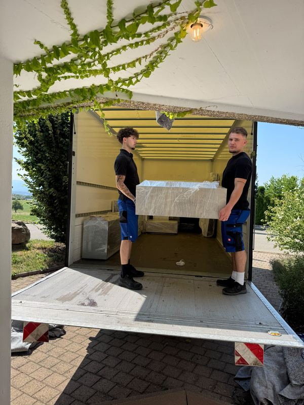 Verpacktes Möbelstück in den Laderaum heben Zwei Kollegen heben ein foliertes Möbelstück von der Ladebordwand in den Laderaum des Umzugswagens